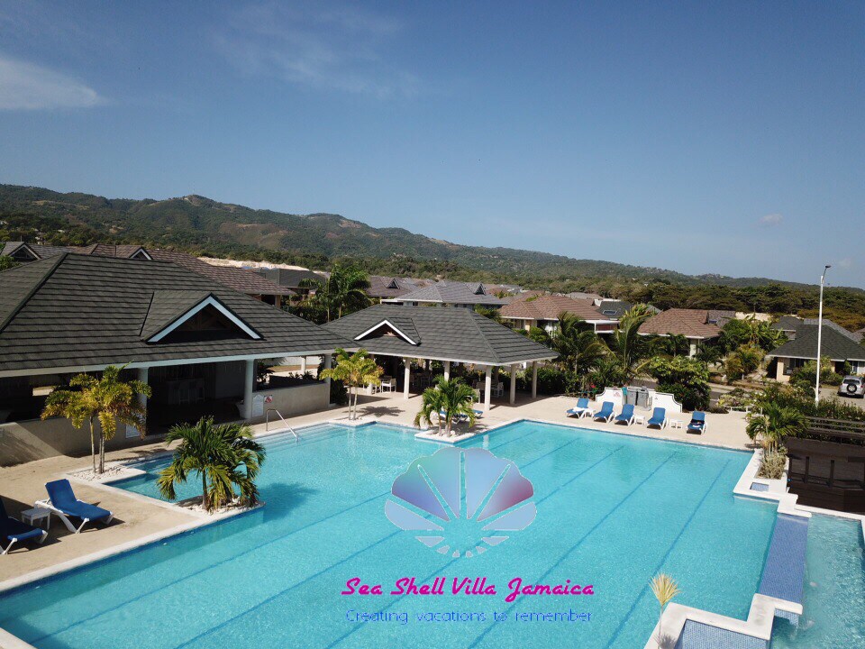 An expansive infinity pool is set within a scenic gated community, featuring numerous lounge chairs around its perimeter. Lush mountains serve as a picturesque backdrop, highlighting the outdoor space's tranquility and inviting environment.