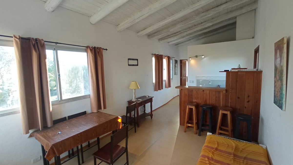 A spacious living area is featured, highlighted by large windows with light brown curtains. A wooden dining table is set with four chairs, while a cozy corner contains a seating area with a colorful blanket. A small kitchenette is visible in the background.