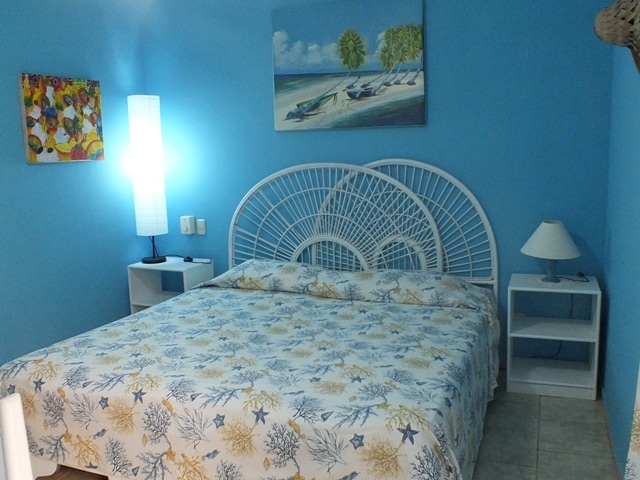 A tranquil sleeping area features a king-sized bed with a patterned bedspread. A lighted floor lamp and a bedside table with a lamp provide soft illumination. Two artworks adorn the blue walls, reflecting coastal themes.