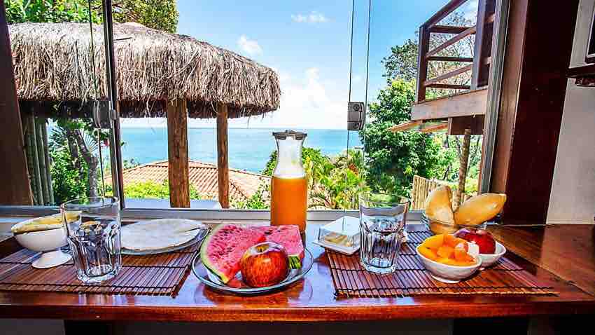 A dining area is presented with a bright view of the ocean through large windows. A table is set with an assortment of fresh fruits, including watermelon and apple, alongside glasses and a pitcher of orange juice, creating an inviting setting for a meal.