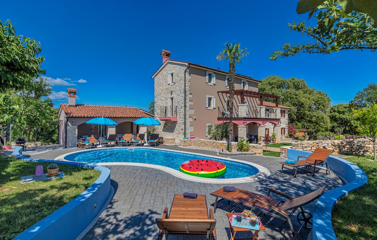 A large swimming pool is surrounded by a grassy area and lounge chairs. The villa's stone exterior is visible in the background, along with vibrant umbrellas providing shade. A colorful inflatable ring adds a playful touch, enhancing the inviting atmosphere of the outdoor space.