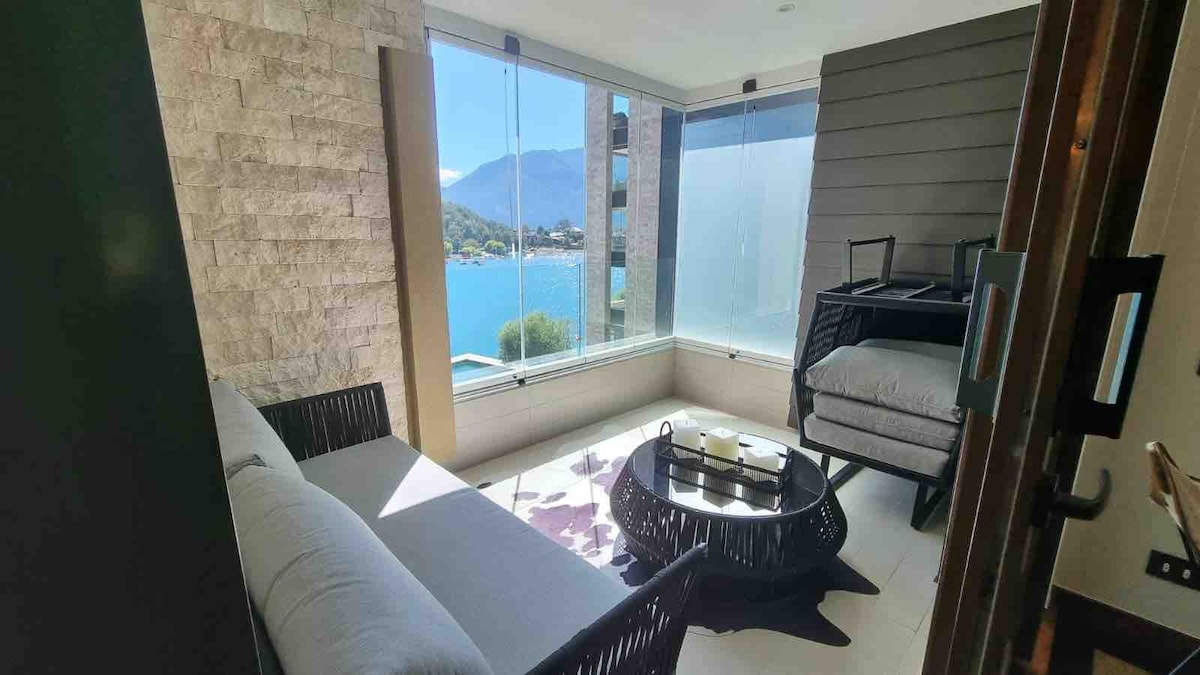 A sunlit sitting area features large windows offering views of the lake and mountains. A comfortable couch is paired with a round coffee table atop a textured area rug, inviting relaxation. Clean lines and natural light create a serene indoor environment.