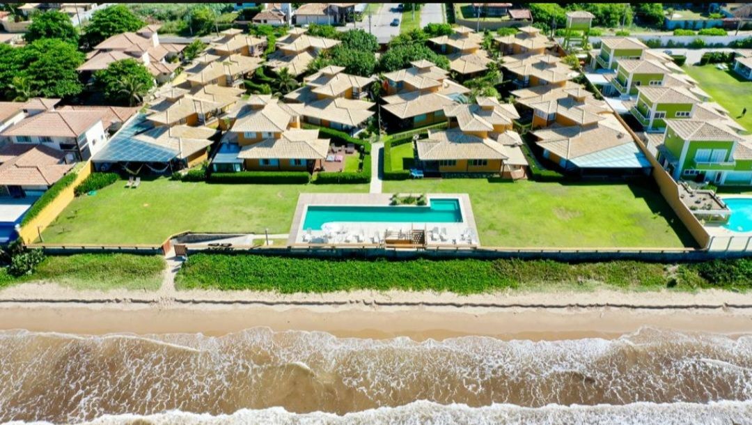 An aerial view captures a well-planned layout of a gated community, featuring multiple houses with sloped roofs surrounded by lush greenery. A panoramic pool is centrally located, bordered by a sun deck, and the sandy beach is visible in the foreground along the coastline.