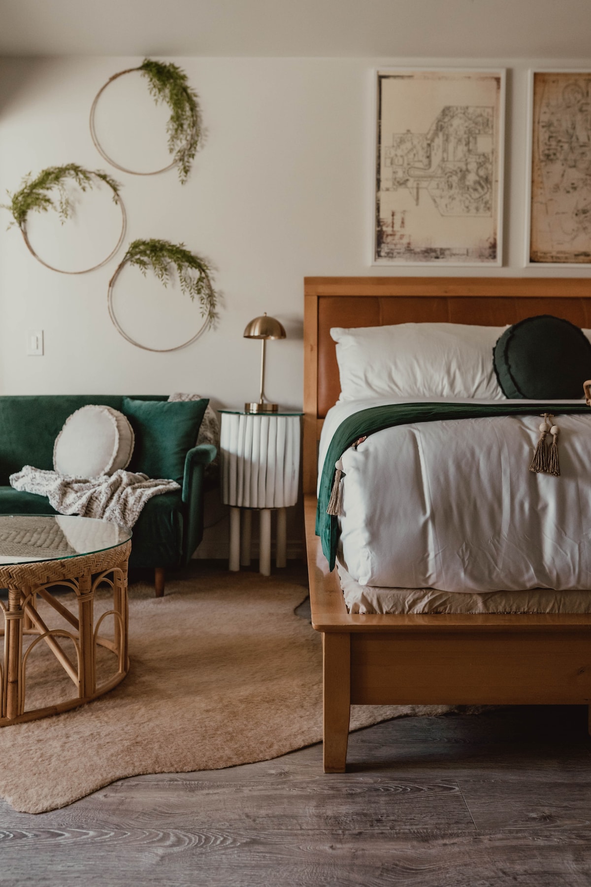 A cozy sleeping area features a spacious bed dressed in neutral linens, accompanied by decorative pillows. A green accent chair adds a pop of color, while a woven coffee table complements the space. Wall decor includes circular greenery arrangements and framed artwork, enhancing the inviting ambiance.