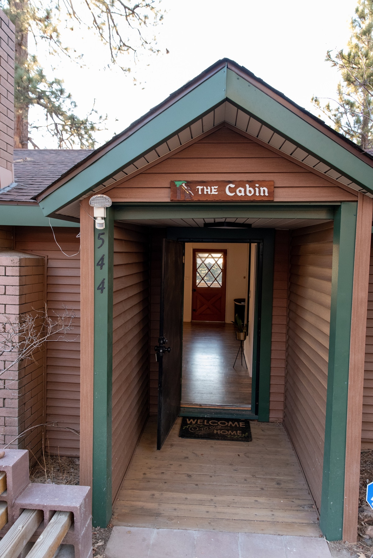 The entrance to the cabin features a brown wooden door that opens into a welcoming foyer. A sign reading 'Welcome' is placed on the ground, and warm, natural tones are observed in the wooden architecture. The inviting entrance provides a glimpse into the spacious interior.