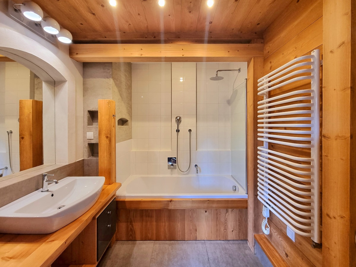 The bathroom features a spacious bathtub with a modern handheld shower attached to the wall. Natural wood accents, including cabinetry and trim, complement the light tile walls. A large mirror above the sink enhances the overall brightness of the space, while a heated towel rack adds functionality.