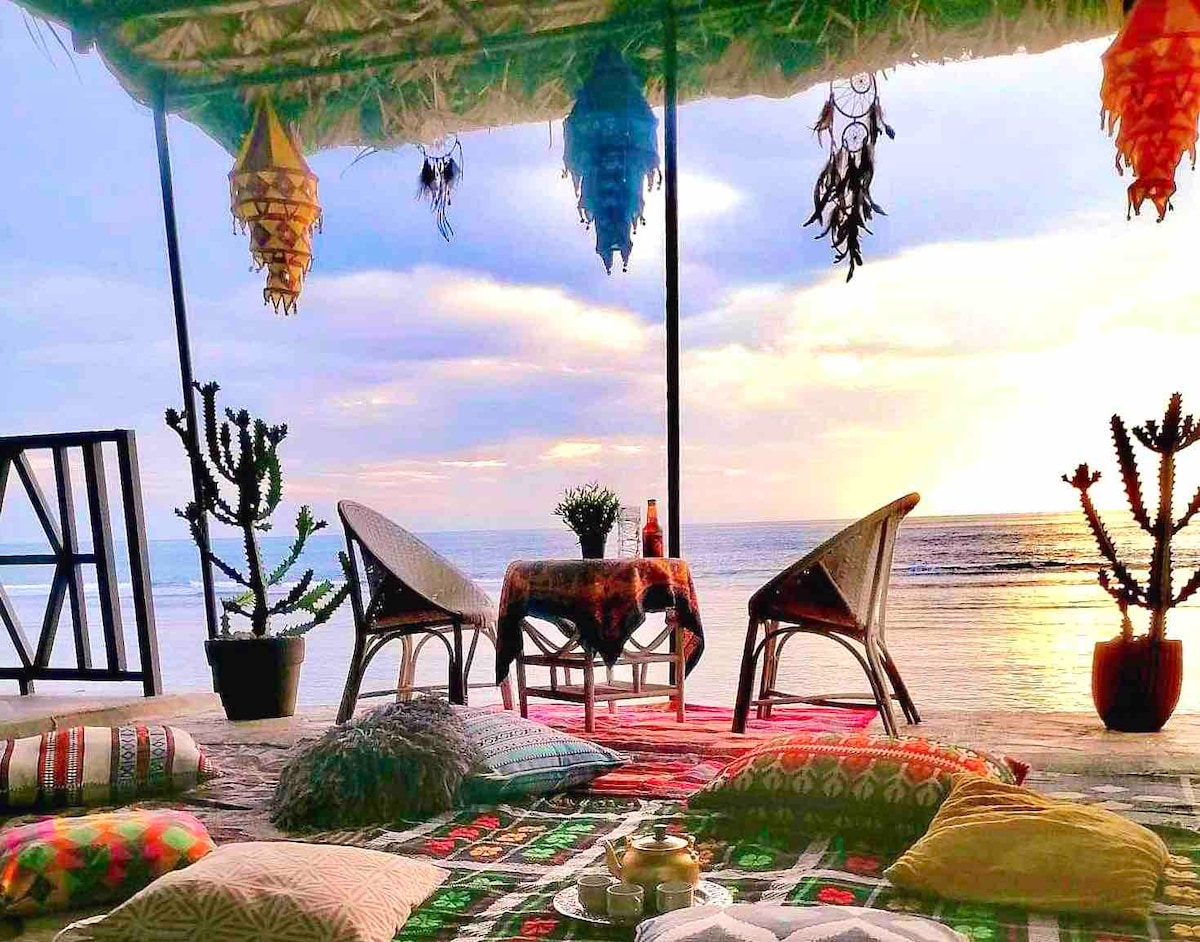 A shaded outdoor seating area features two woven chairs and a low table adorned with a decorative tablecloth. Colorful pillows are scattered across a patterned mat, surrounded by potted plants. The backdrop includes a serene view of the sea and a sunset sky, enhancing the tranquil setting.
