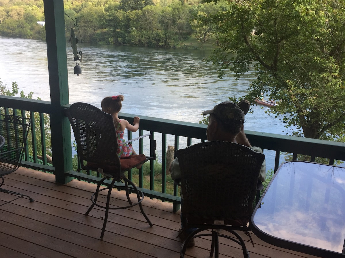 A covered deck overlooks the peaceful river, with a young child standing beside a seated adult. Two bar-style chairs and a small table are positioned on the wooden deck, providing a space for relaxation and views of the lush greenery along the water.