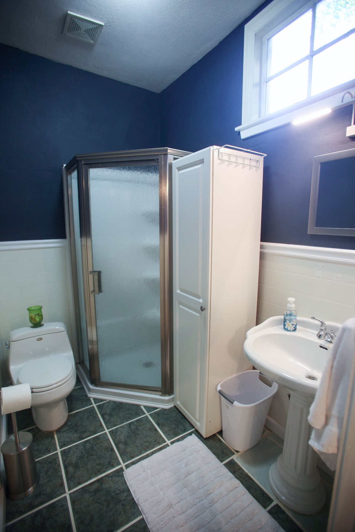A compact bathroom features a glass-enclosed standing shower, a toilet, and a pedestal sink. A large window allows natural light to enter, while white and navy blue walls create a clean look. A light-colored rug lies on the tiled floor, complementing the overall design.