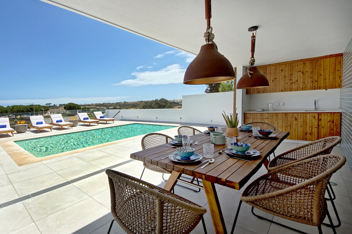 An outdoor dining area features a wooden table surrounded by woven chairs, positioned beside a pool. The space is complemented by hanging pendant lights and a kitchenette. Sun loungers are arranged near the pool, offering views of the surrounding landscape under a clear sky.