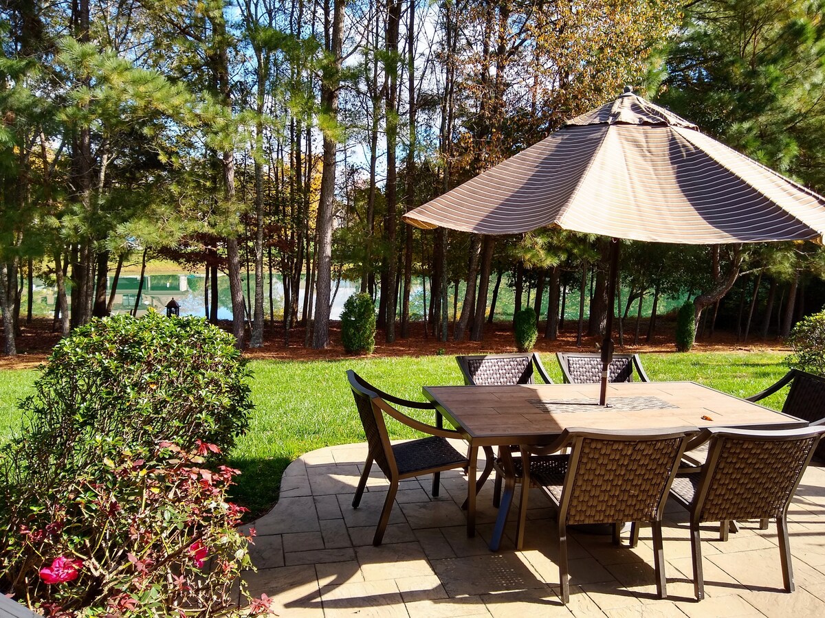 An outdoor dining area is featured, with a large table surrounded by wicker chairs. A shaded umbrella provides cover, while landscaping with shrubs and a verdant lawn leads to a serene lake in the background, surrounded by trees.