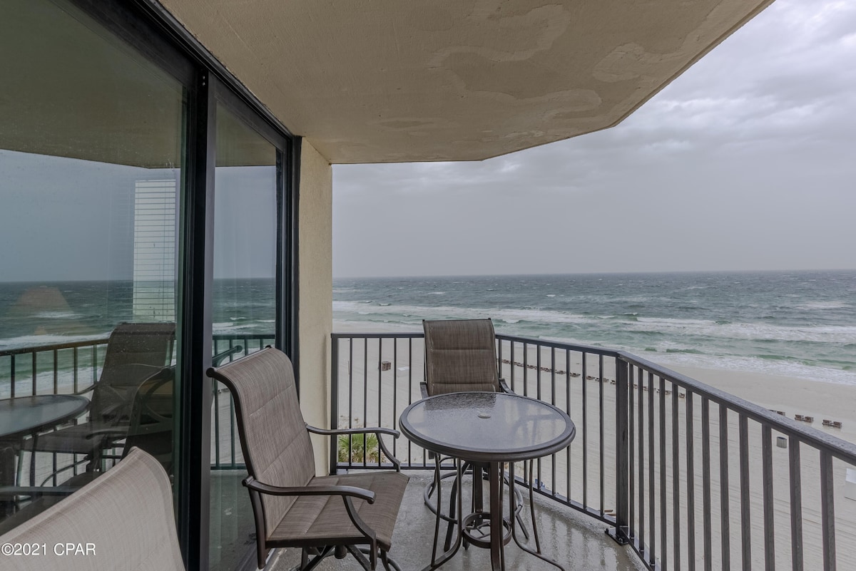 A coastal balcony features a round table with two chairs, overlooking the ocean waves. Glass doors provide access to the living space, while the expansive view includes the beach and shoreline. The scene is accented by a cloudy sky, hinting at the ocean's ambiance.