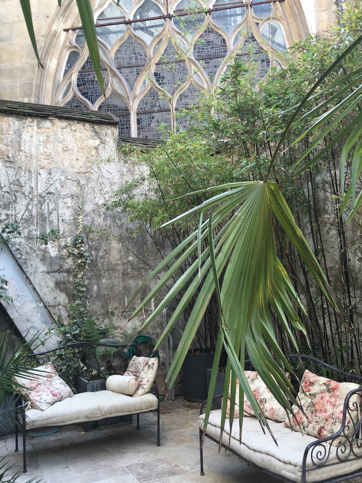 A serene outdoor terrace features two lounge chairs with cream cushions and floral accents. Lush greenery surrounds the space, with tall plants and a backdrop of an elegantly designed historic stone wall and large gothic window, providing an atmosphere of tranquility.