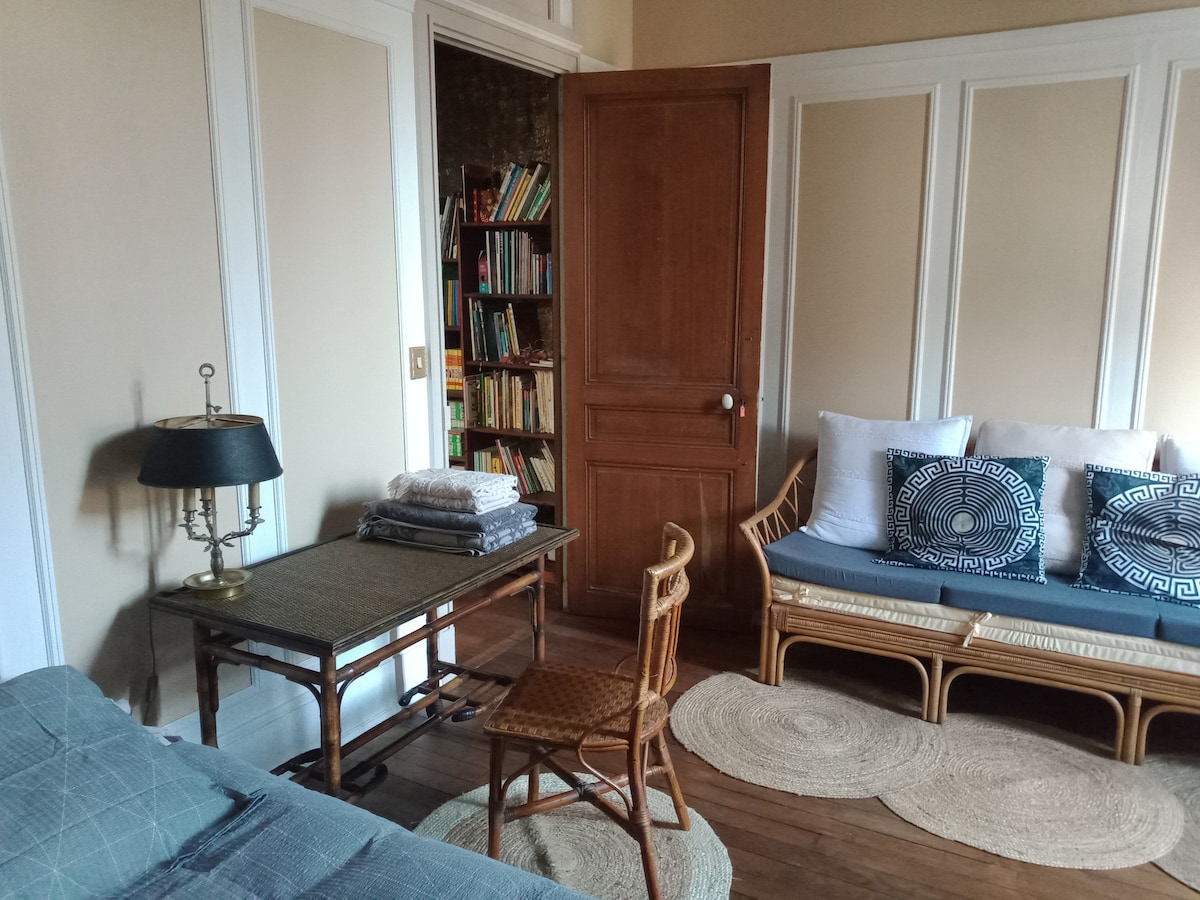 A cozy room features a wooden table with a lamp, and a comfortable seating area with a blue sofa adorned with patterned cushions. A stack of towels is neatly arranged on the table. A charming bookshelf can be seen in the background, enhancing the room's warm feel.