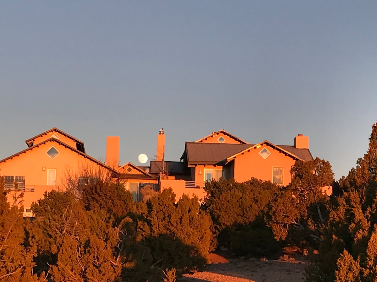 A serene desert landscape is captured at sunset, highlighting a large adobe-style house with multiple gabled roofs and distinctive windows. Surrounding juniper trees reflect a warm glow from the setting sun, creating a tranquil ambiance against the evening sky.