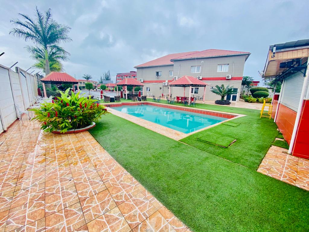 A well-maintained outdoor area features a crystal-clear swimming pool surrounded by landscaped greenery. Comfortable lounge chairs are arranged near the pool, while several shaded seating areas are available. Two-story buildings with red roofs are situated in the background, enhancing the serene atmosphere.