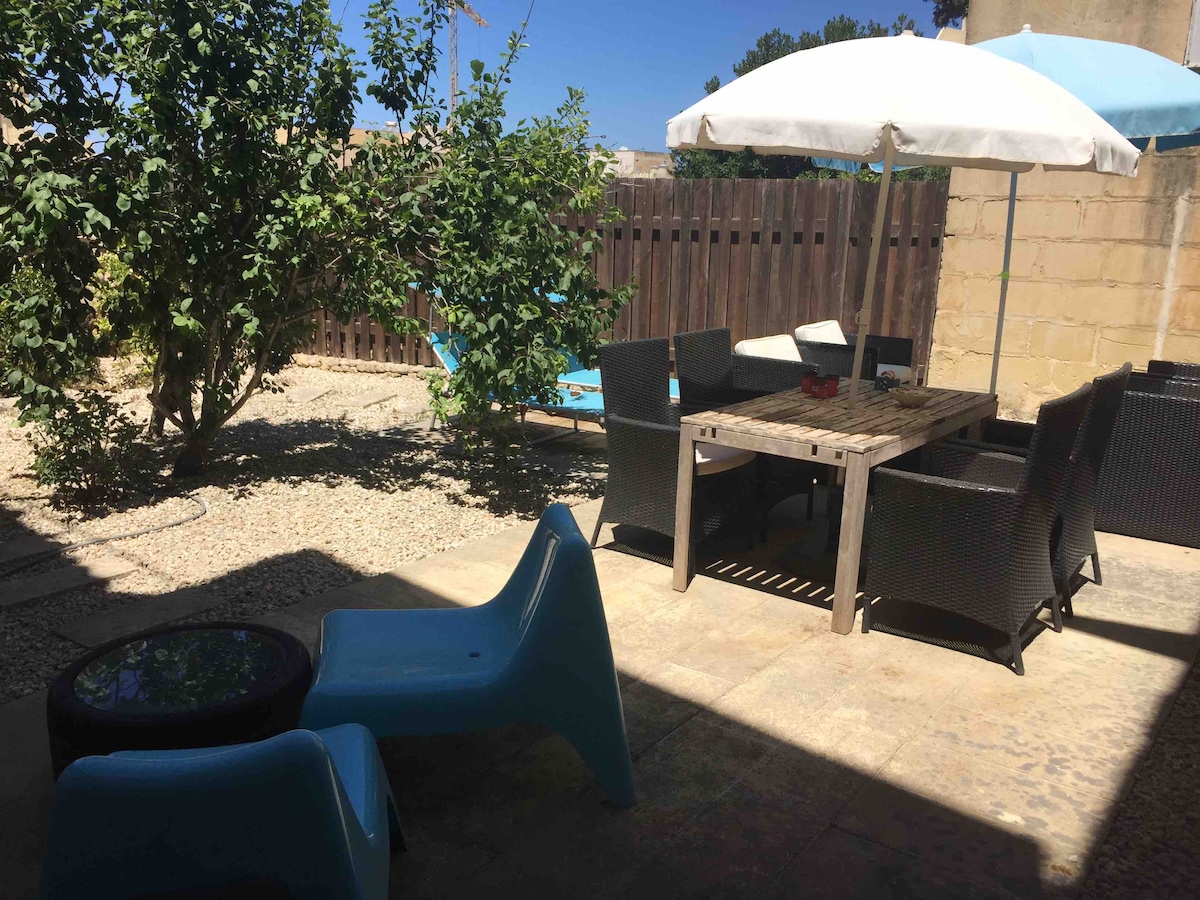 An outdoor garden area features a wooden dining table surrounded by dark wicker chairs. A large umbrella provides shade, while a pair of blue lounge chairs rests in a gravel space. Lush green trees add a natural touch to the inviting setting.