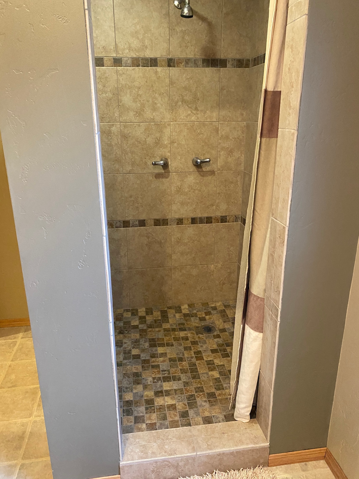 A spacious shower area is depicted with tiled walls and a textured floor. Two chrome shower heads are mounted on the wall, and a neutral-toned curtain is partially drawn. The earthy tones and decorative tile give a natural feel to the space.