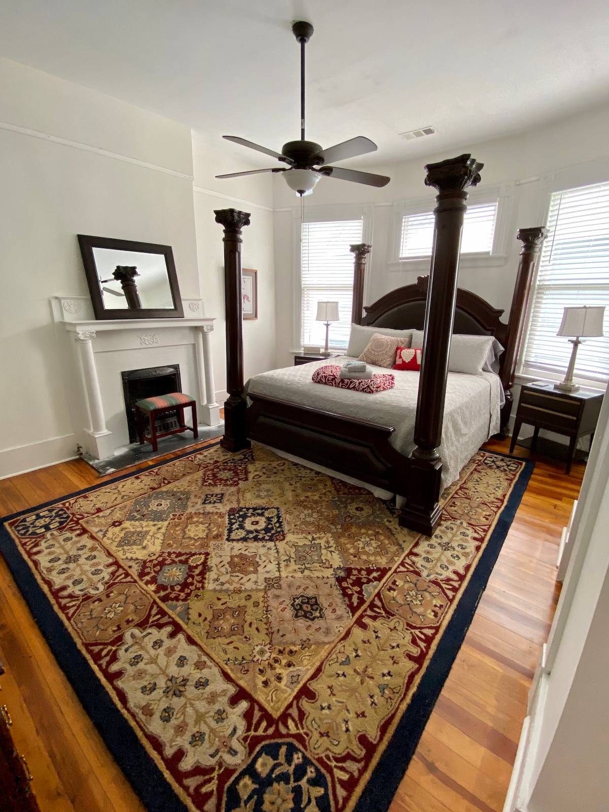 A well-appointed bedroom features a king-size bed with plush bedding against a backdrop of large windows. A decorative area rug enhances the space, while a traditional fireplace adds character. Two bedside tables with lamps complete the inviting atmosphere.