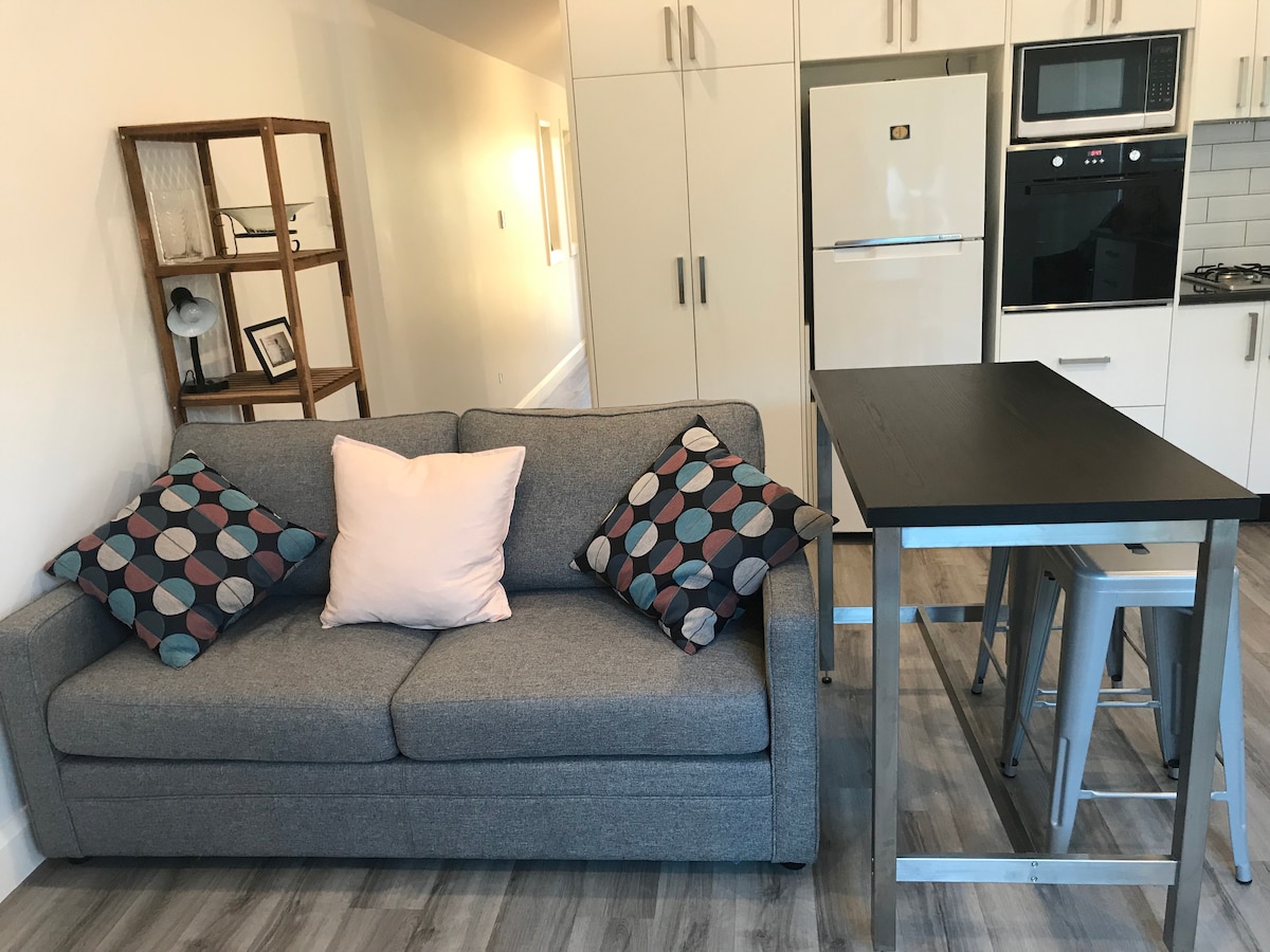A modern living area includes a gray sofa adorned with patterned cushions and a light pink throw pillow. A sleek dining table with metallic stools is positioned nearby, complemented by white cabinetry and stainless steel appliances in the background.