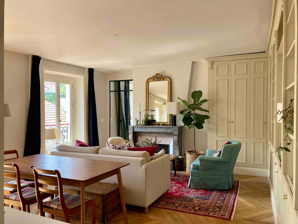 A spacious living area is highlighted by natural light streaming through large windows. A comfortable sofa and armchair are positioned around a central coffee table, with a stylish rug adding color to the hardwood floor. A decorative fireplace and a large mirror enhance the inviting ambiance.