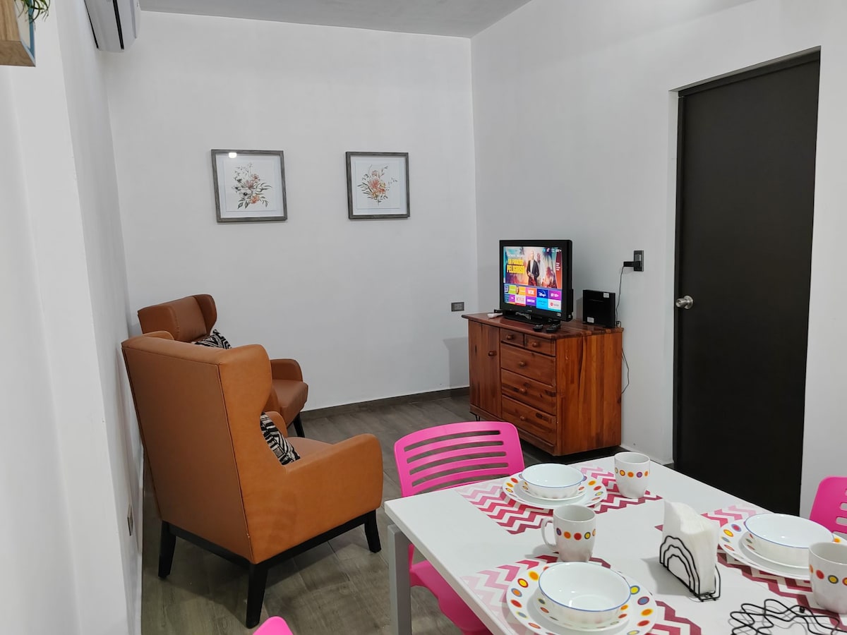 A cozy living area is present, featuring two orange armchairs positioned near a wooden television stand. A television displays vibrant images. A white dining table is set with decorative plates and utensils, complemented by pink chairs. Framed floral artwork adorns the walls.