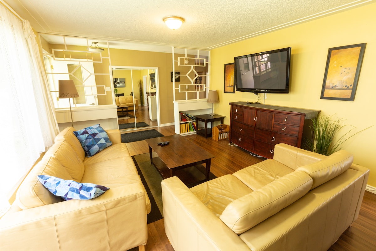 The living area features two comfortable, light-colored sofas facing a wooden coffee table. A large flat-screen TV is mounted on the wall opposite the seating. Soft natural light filters through sheer curtains, and decorative artwork is displayed on the walls.