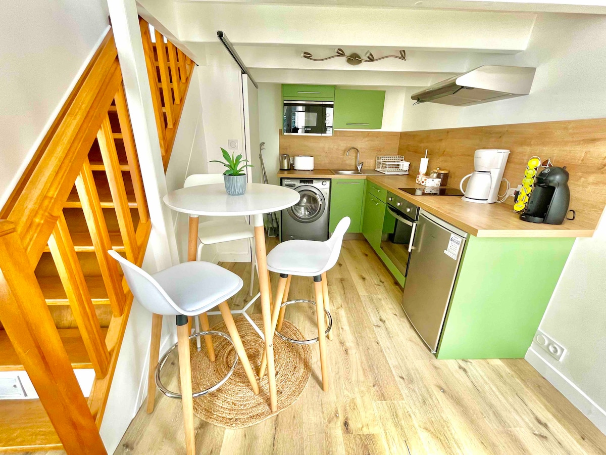 A compact kitchen area features modern appliances, including a washing machine, and is accentuated by green cabinetry. A small round dining table with three high stools is included, along with wooden accents from the stairs. Natural light illuminates the space.
