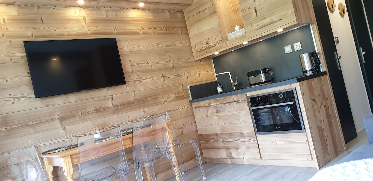 A modern kitchen area is presented with wooden cabinetry and a sleek countertop. It features a built-in oven, stovetop, and essential appliances, including a kettle and coffee maker. A television hangs above the countertop, while transparent chairs surround a dining area, creating a clean and functional space.