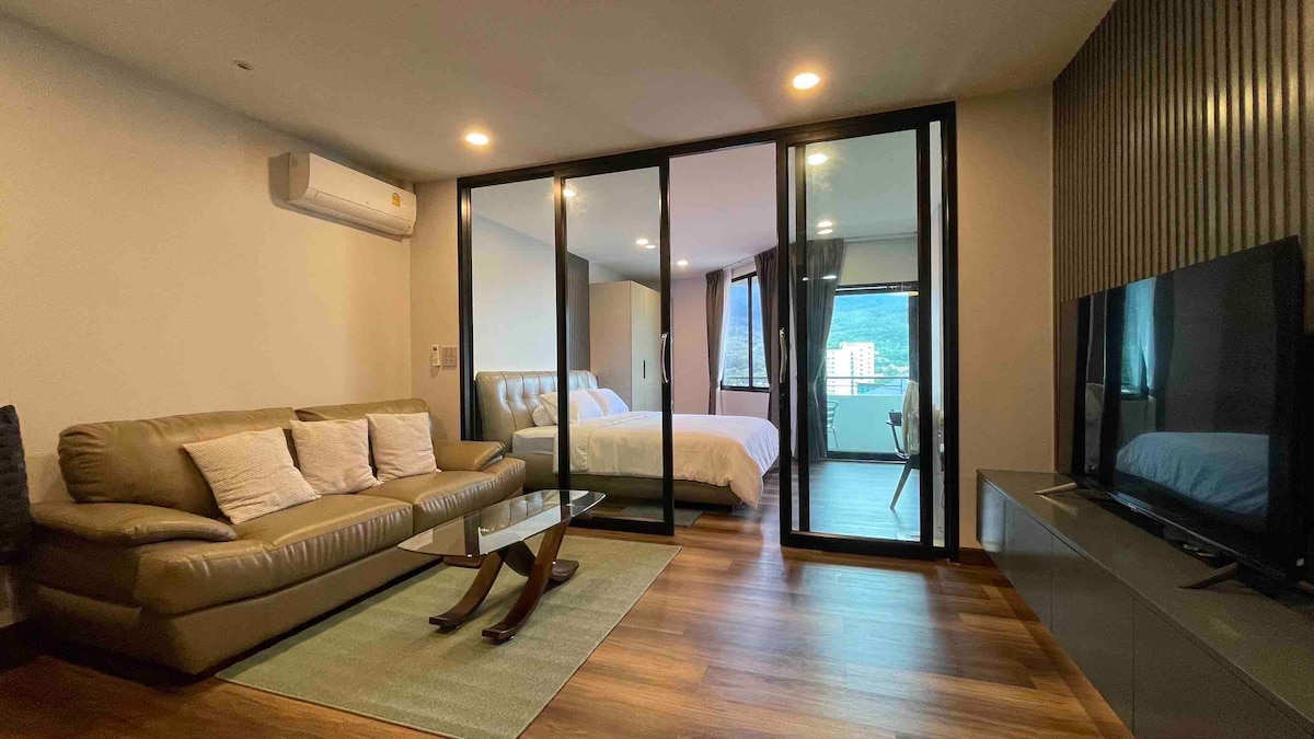 A spacious living area features a comfortable sofa with several throw pillows, complemented by a glass coffee table. Large glass doors offer a view of the bedroom, furnished with a broad bed and natural light streaming in, while a television stands on a low cabinet.