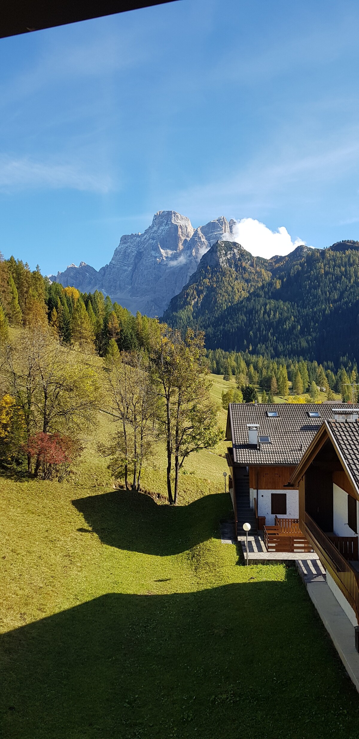 A panoramic view of the surrounding mountains showcases lush greenery and distant peaks under a clear blue sky. The image captures a blend of open hillside and wooden structures, with shadows cast by the trees and buildings, creating a serene outdoor atmosphere.