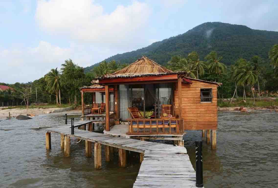 A charming wooden villa is situated on water, accessed by a sturdy wooden pier. The structure features a thatched roof and large windows, inviting natural light. Surrounding palm trees enhance the tropical ambiance, with distant mountains visible in the background.