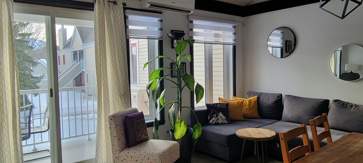 A bright living area is depicted, featuring a spacious sectional couch adorned with pillows, a wooden coffee table, and a large indoor plant. Natural light filters through sliding glass doors that open to a balcony, with modern mirrors adding depth to the decor.