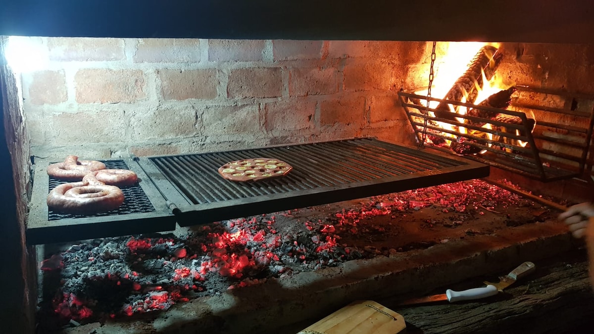 A traditional outdoor grill is shown, featuring a charcoal fire with glowing embers. Sausages are placed on the grill, while a flatbread rests nearby. The surrounding brick wall is illuminated by soft light, creating a warm and inviting cooking space.