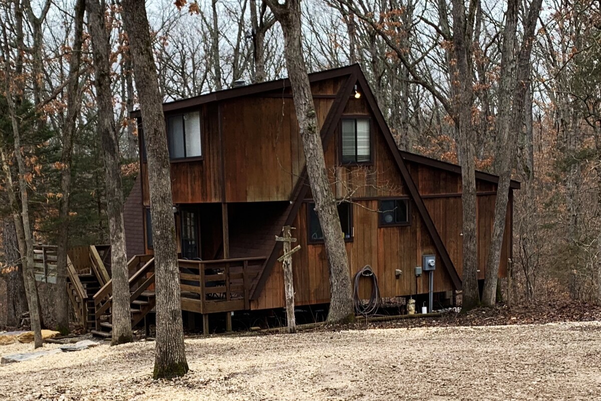 The cabin is set in a wooded environment, showcasing a rustic design with warm wooden siding. The structure features multiple windows and a slanted roof. A wooden deck can be seen at the entrance, surrounded by trees, creating a private and tranquil atmosphere.