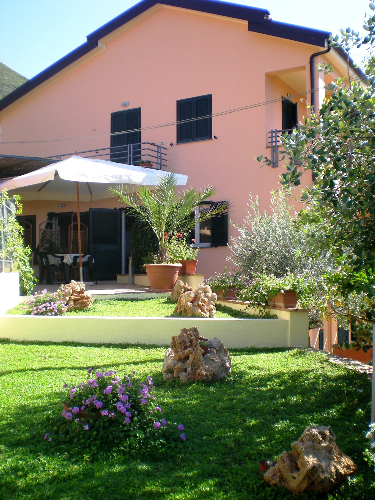 A charming pink building features a well-maintained garden with green grass and various plants. Outdoor seating is provided under an umbrella, with stone decorations adding character. The entrance is visible, welcoming guests to the inviting space.