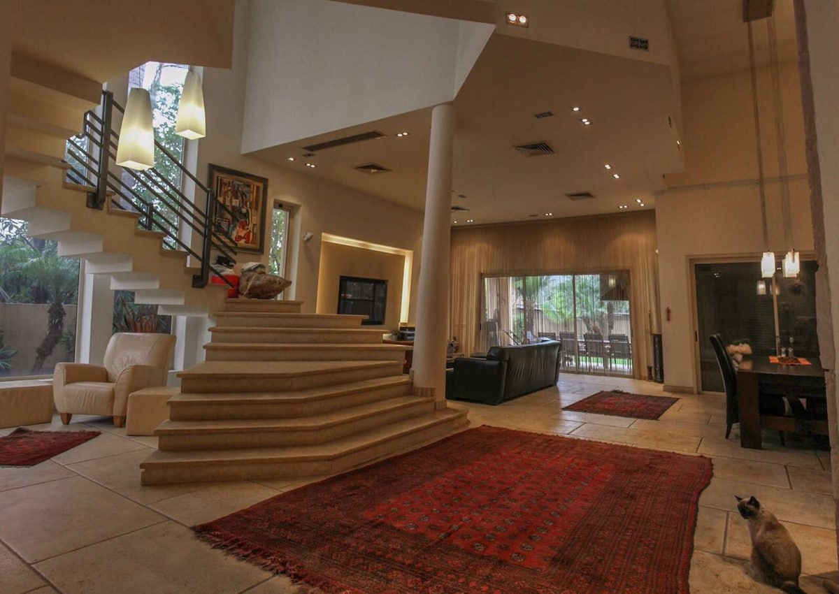 The interior space features a spacious living area with high ceilings and natural light flowing through large windows. A striking staircase leads upstairs. Elegant furnishings include a casual seating area and a dining table, complemented by decorative rugs and subtle artwork on the walls.