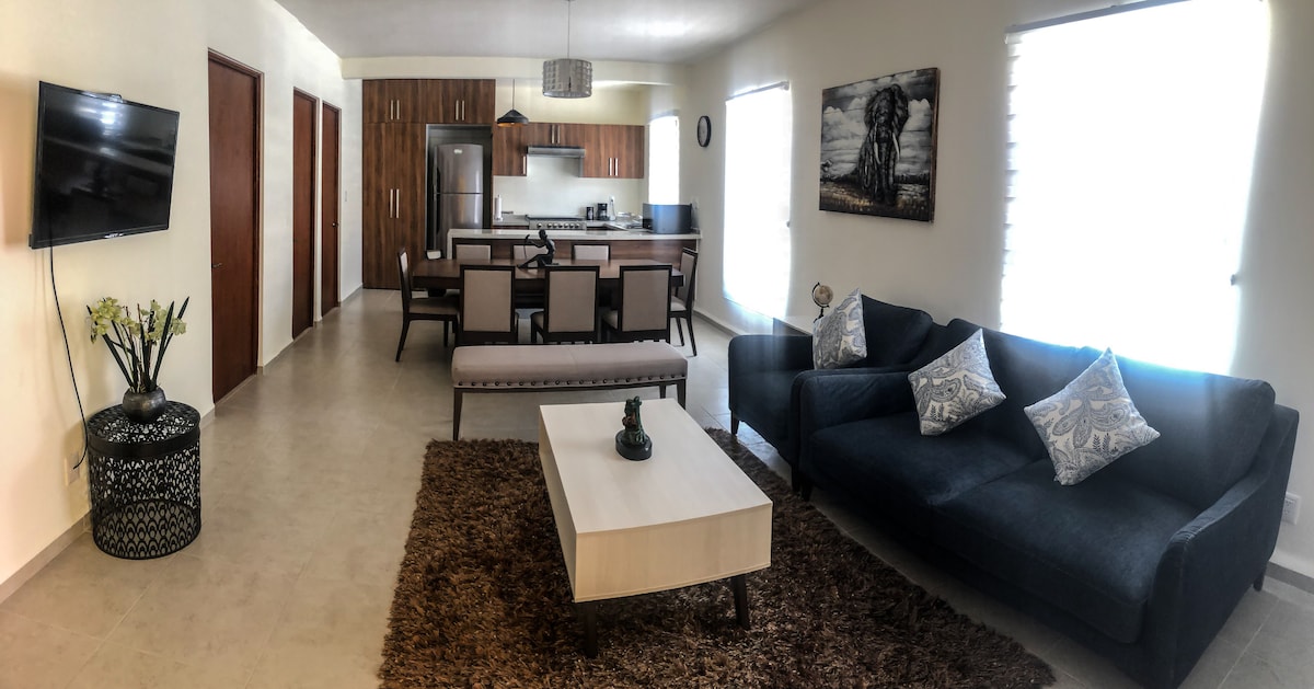 A modern living space features a stylish dark sofa adorned with decorative pillows, a light wooden coffee table, and a dining area with six chairs. A mounted TV is visible on the wall, and natural light enters through large windows with light treatments. Decorative artwork complements the design.