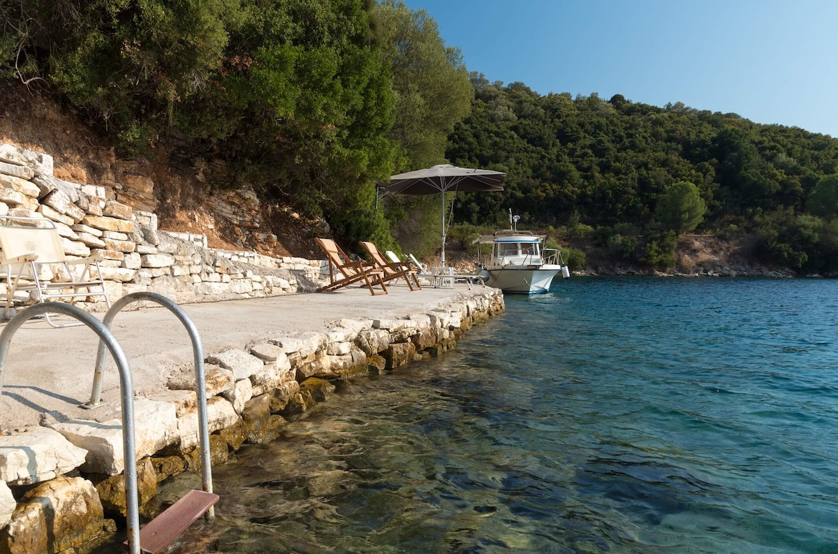 A tranquil waterfront area features a stone pathway leading to clear turquoise waters. Sun loungers are arranged under a large umbrella on the dock, which provides access to the sea. A small boat is visible nearby, surrounded by lush greenery along the shoreline.