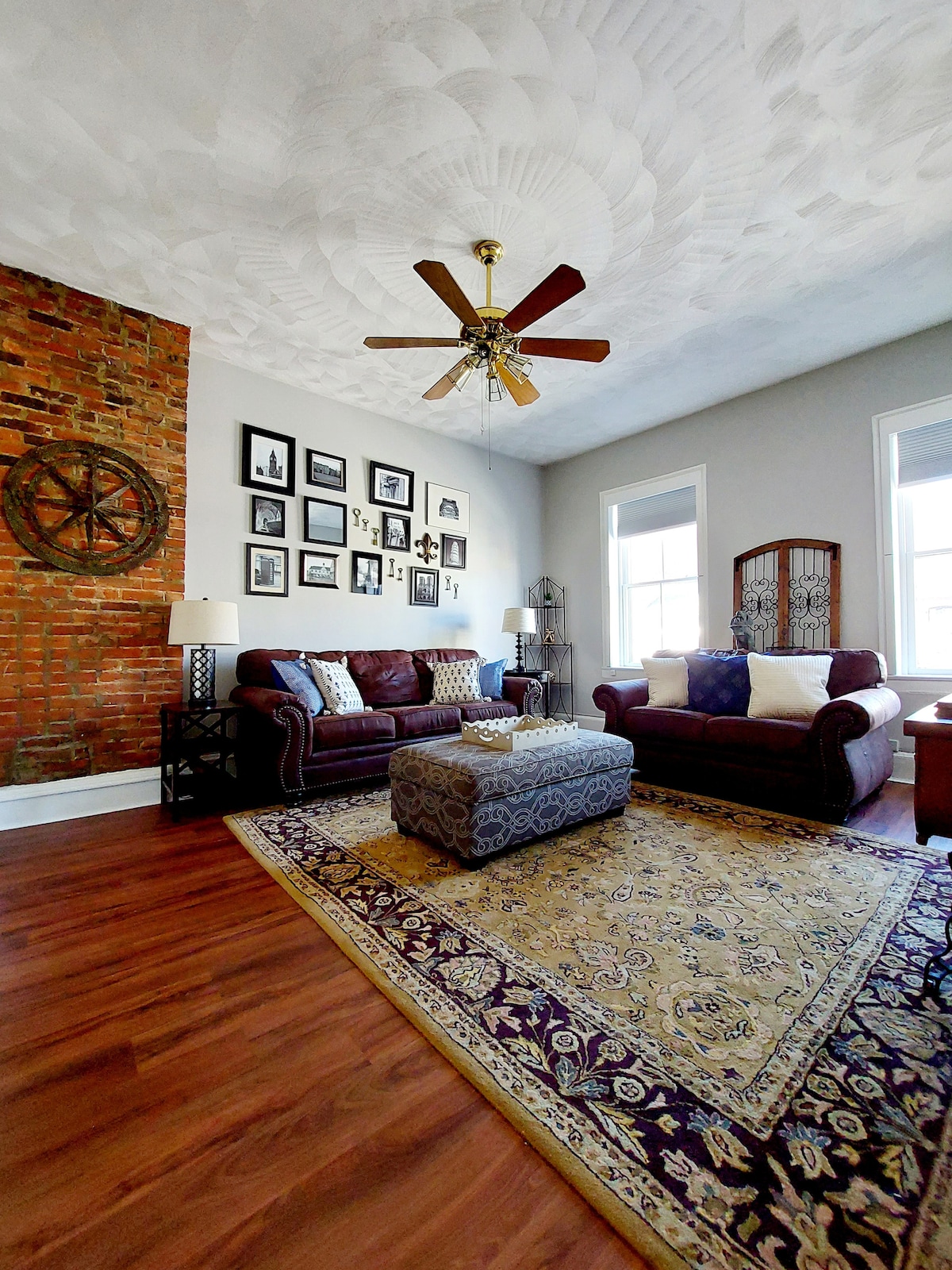 The living room features a mixture of comfortable furniture, including a dark leather sofa and armchair. A decorative area rug lies under a coffee table, while a ceiling fan hangs above. A brick accent wall displays framed photographs, providing visual interest and a cozy atmosphere.