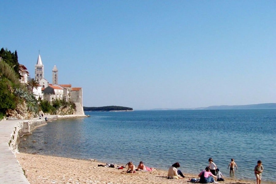 A sandy beach stretches along the shoreline, where several individuals relax in the sun. The calm blue water gently laps against the shore, with a quaint coastal village visible in the background, featuring traditional buildings and a church tower.