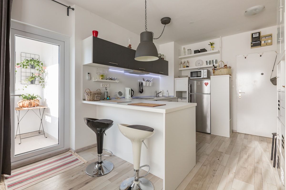 A modern kitchen area features a sleek island with two bar stools. Dark cabinetry contrasts with light countertops. Shelves display various kitchen items. A doorway to a balcony is visible, along with a view of greenery. The floor showcases wood-like tiles, enhancing the fresh ambiance.