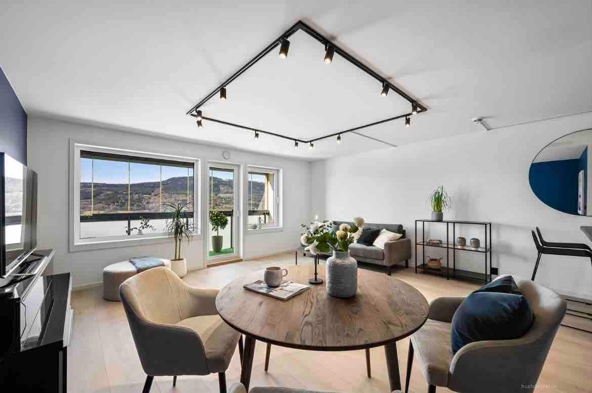 A modern living space is highlighted by natural light from large windows, showcasing a round wooden table surrounded by comfortable chairs. A sleek gray sofa and decorative plants add warmth to the neutral-toned environment, while stylish track lighting enhances the overall contemporary aesthetic.