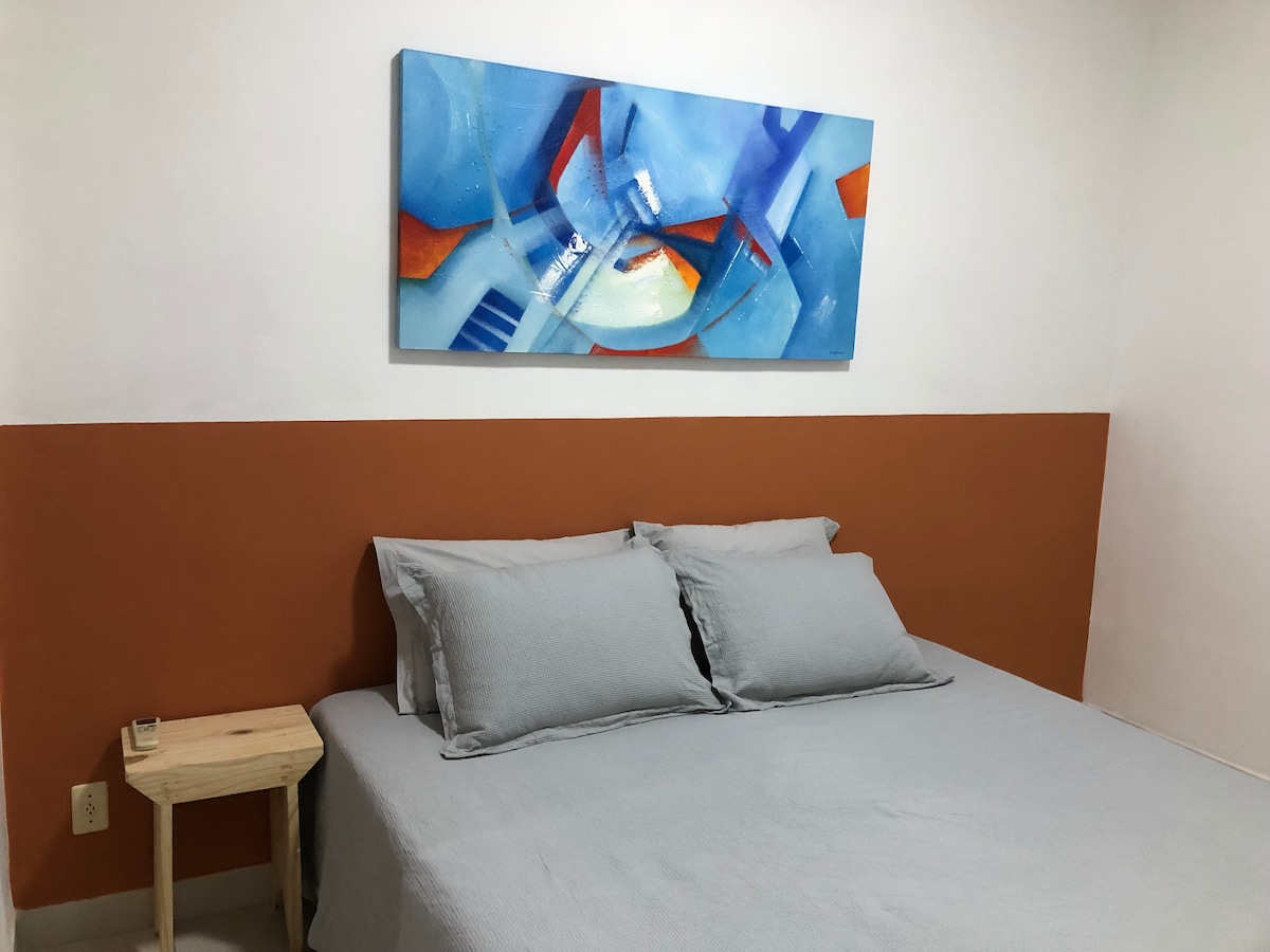 A spacious bed with light grey bedding is positioned against a white wall. Above the bed, an abstract painting featuring blue, orange, and white tones adds a splash of color. A small wooden bedside table is placed next to the bed.