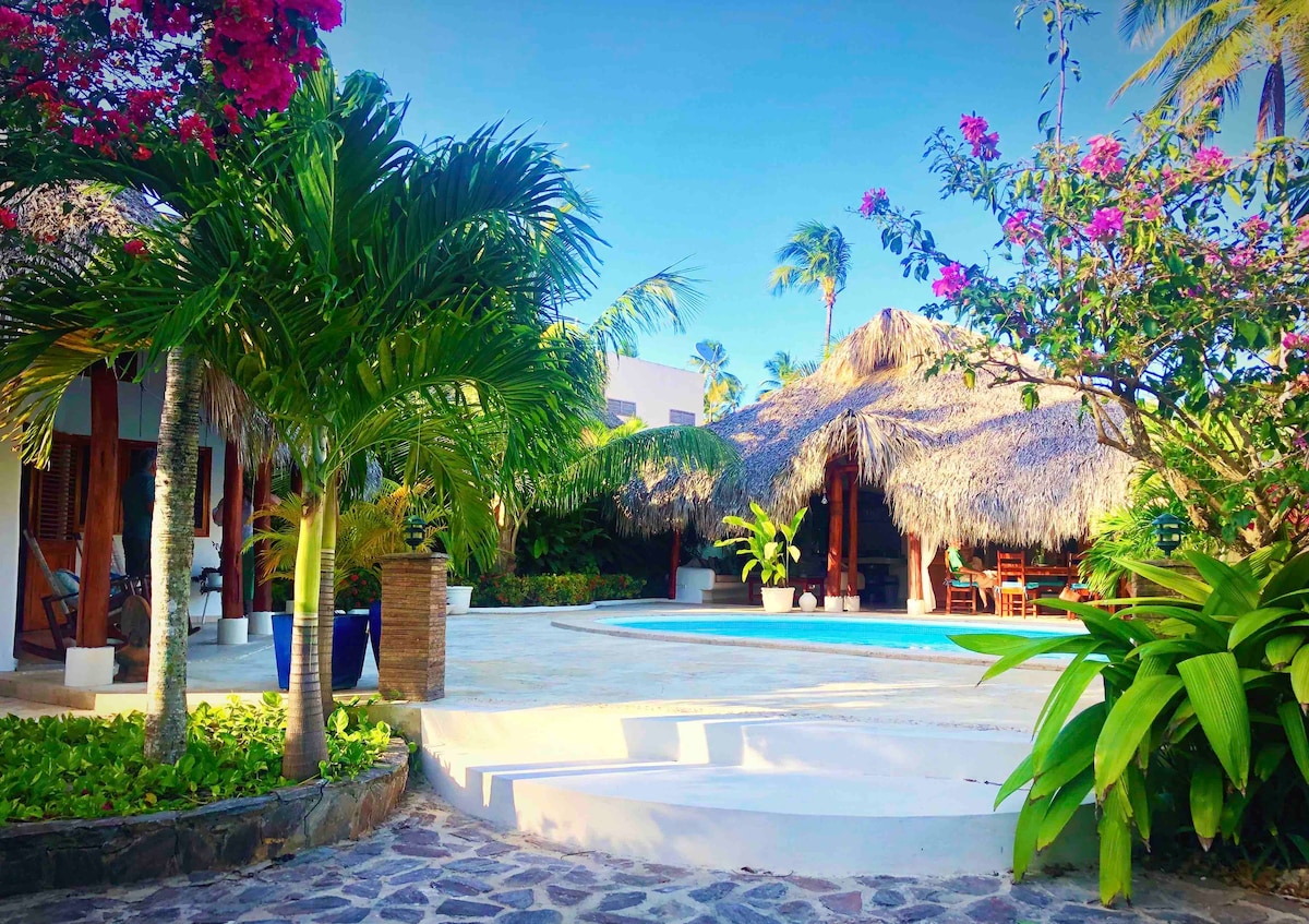 A thatched-roof porch provides shaded dining and seating areas near a large saltwater pool. Lush palm trees and flowering plants frame the scene, complementing the bright blue sky. The pool area features smooth stone pathways leading to the various living spaces.