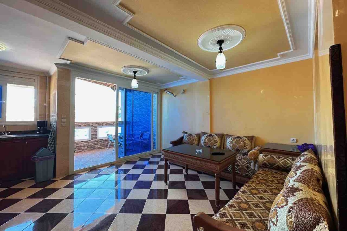An inviting living area features a checkerboard tile floor and elegant ceiling details. A patterned sofa set, complemented by a wooden coffee table, is positioned near large glass doors that open to a balcony, allowing natural light to brighten the space.