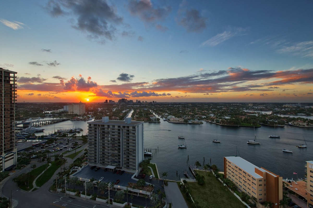 A panoramic view captures a sunset over the Intracoastal Waterway, with vibrant hues of orange, pink, and purple reflecting on the water. Marina boats dot the scene, framed by waterfront properties and lush greenery, enhancing the serene ambiance of the setting.