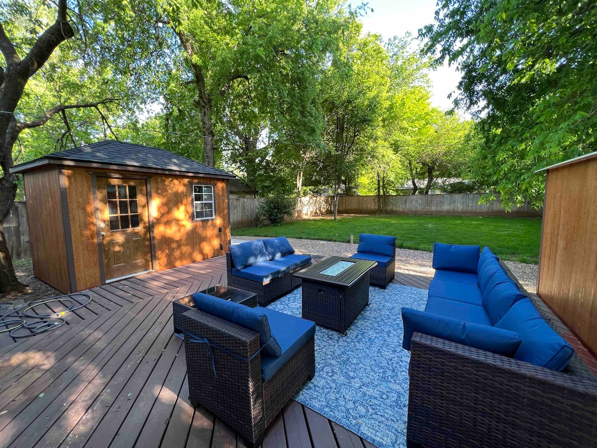 A spacious outdoor patio is highlighted by comfortable blue seating and a table situated on a patterned rug. Surrounded by lush greenery, a wooden shed is visible in the background, creating a relaxed atmosphere for social gatherings.