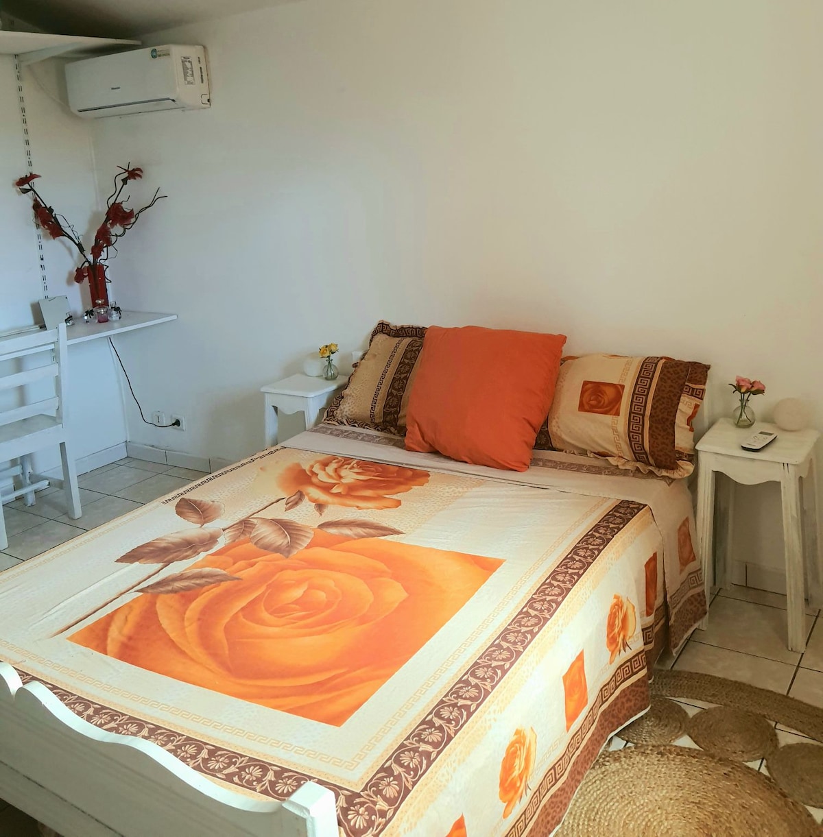 A comfortable sleeping area features a spacious bed adorned with a floral-patterned blanket and matching decorative pillows. Two nightstands are positioned on either side, each with a small vase. Natural light enters through a nearby window, while air conditioning provides climate control.