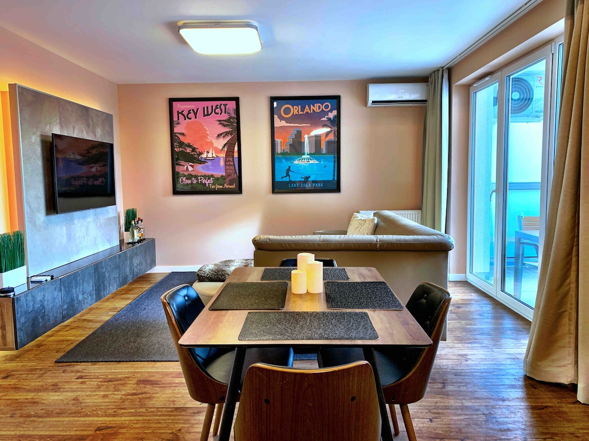 An open living area features a modern sectional sofa, a large flat-screen TV mounted on the wall, and a wooden dining table with four chairs. Two travel-themed posters adorn the wall, and large glass doors provide access to the balcony.
